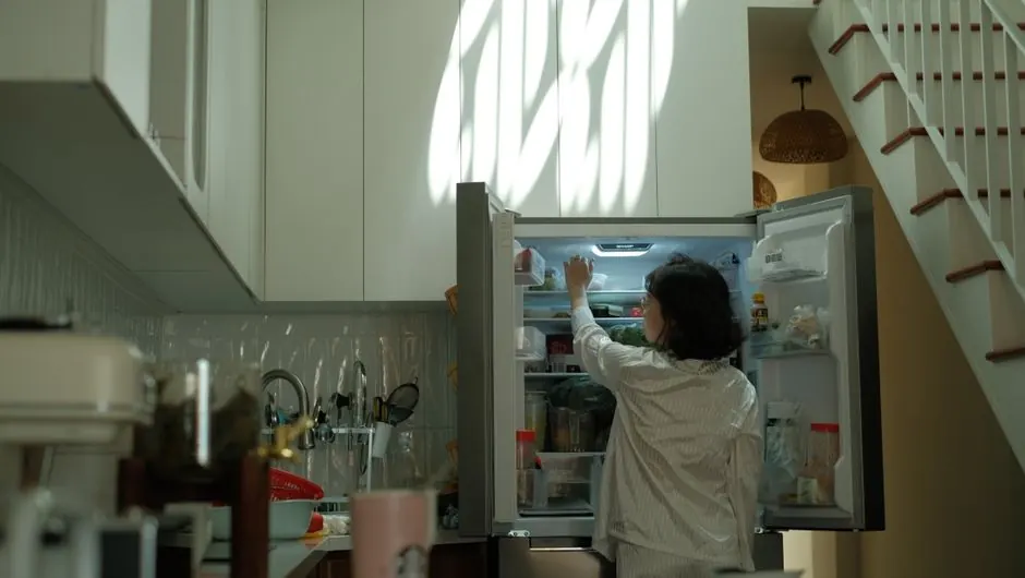 A woman in pajamas checks the refrigerator in a warmly lit cozy kitchen.